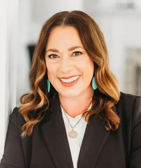 Headshot of Meredith LeConey smiling warmly, wearing a black blazer, white blouse, teal earrings, and a layered gold necklace against a bright neutral background