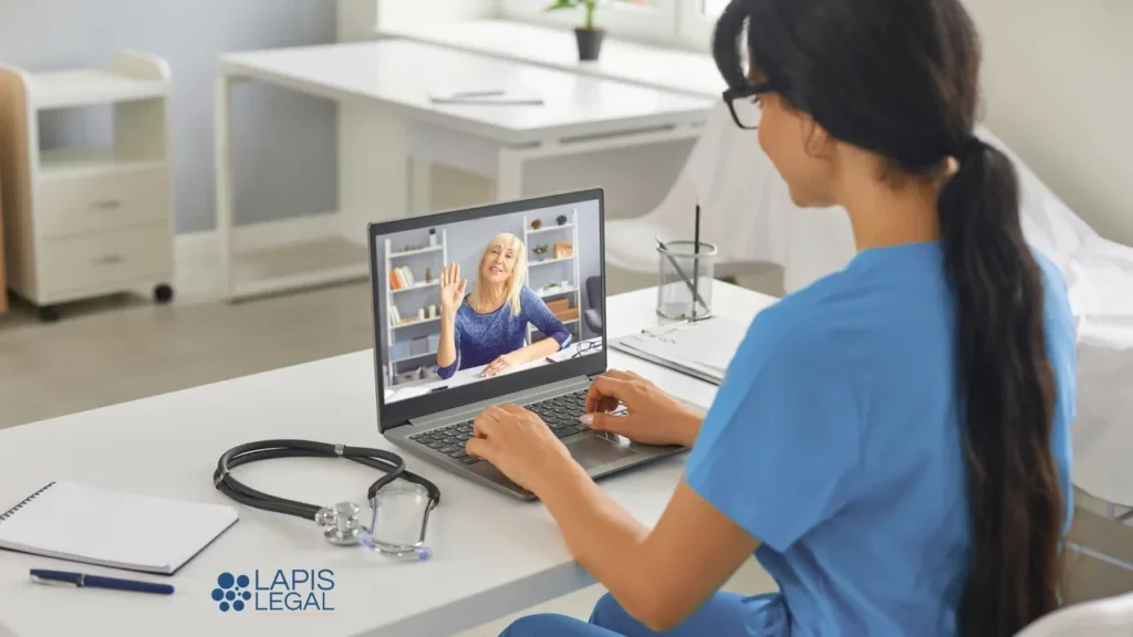 Healthcare professional in blue scrubs on a video call with a patient on a laptop, with a stethoscope on the desk nearby