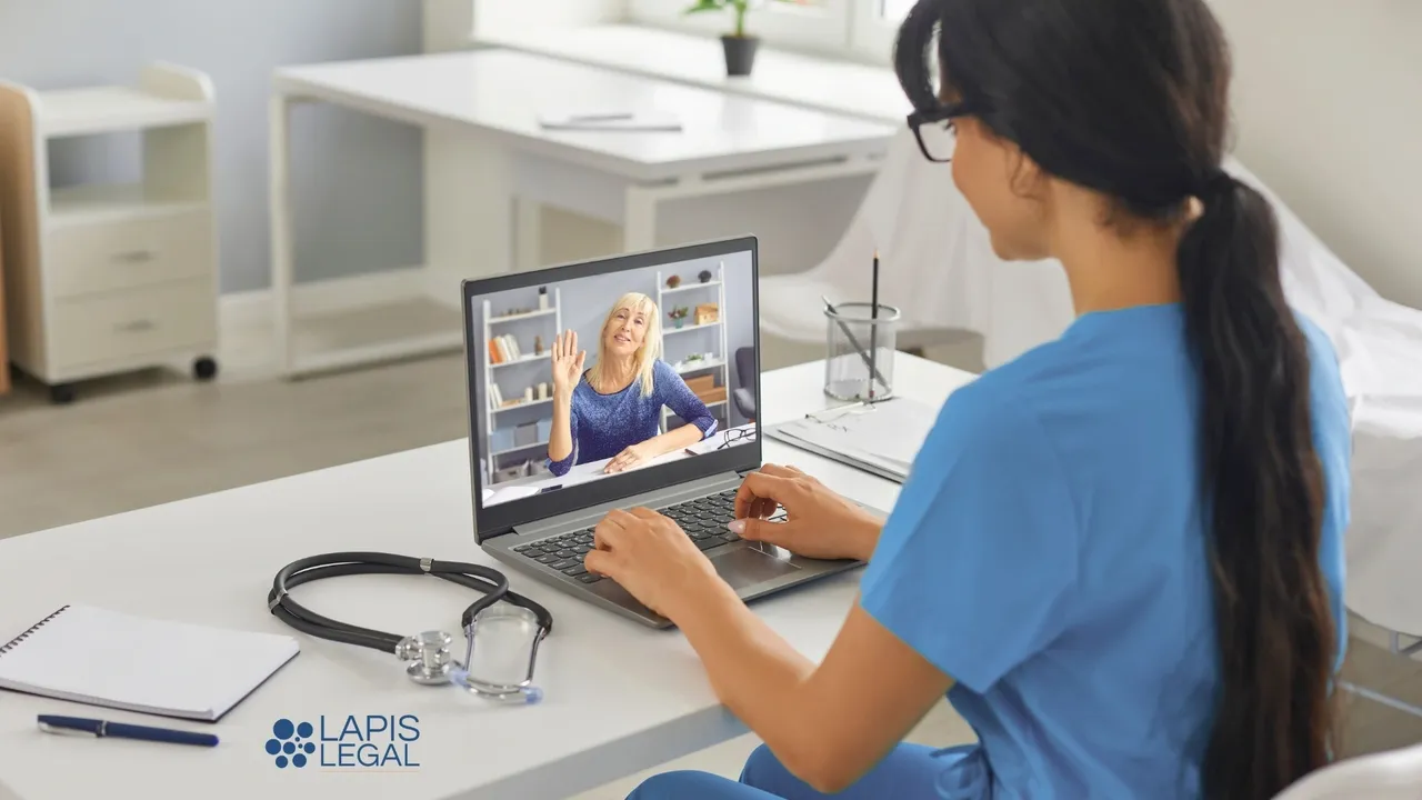 Healthcare professional in blue scrubs on a video call with a patient on a laptop, with a stethoscope on the desk nearby