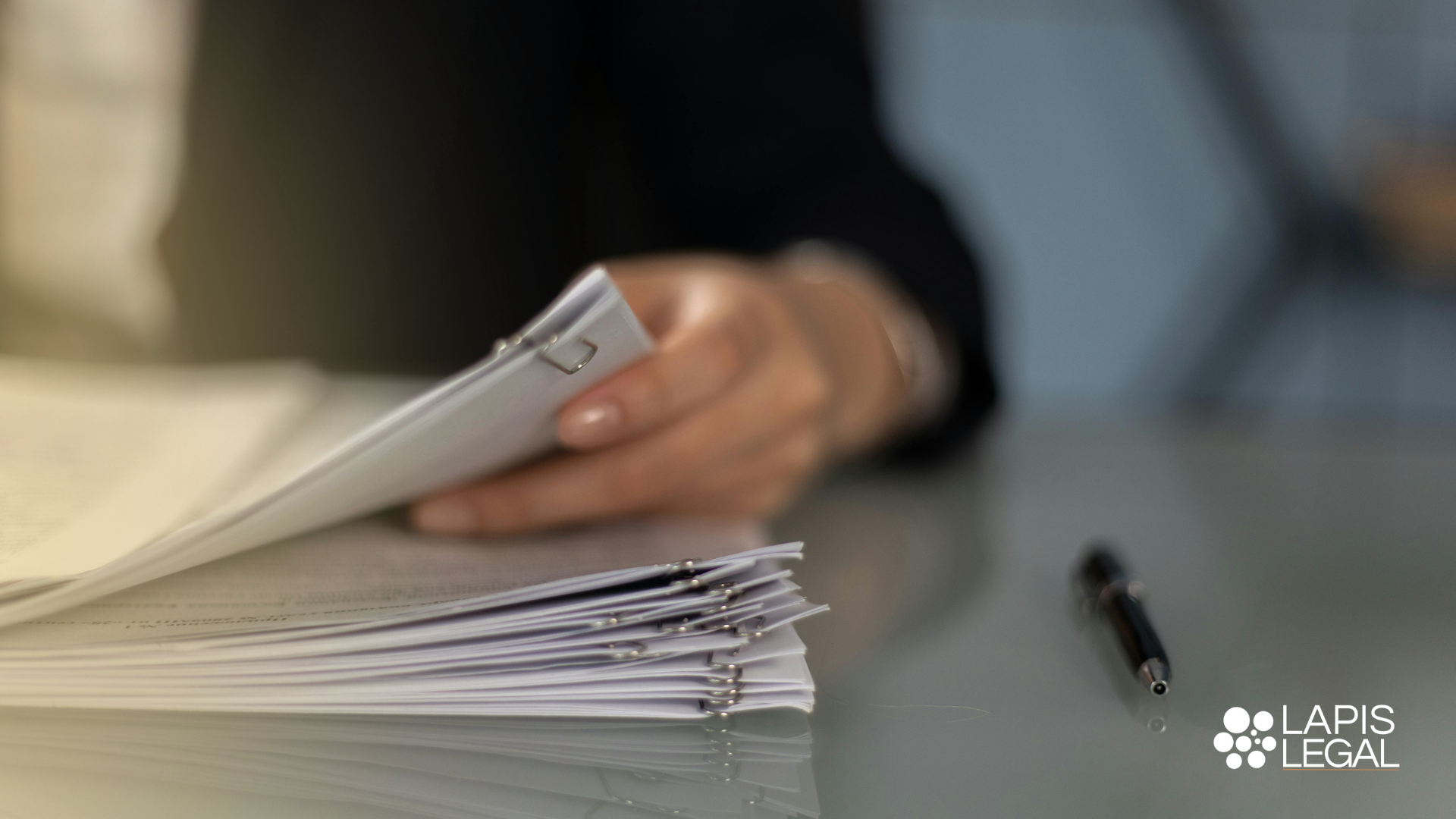 Person's hand holding a stack of clipped documents on a desk with a pen nearby